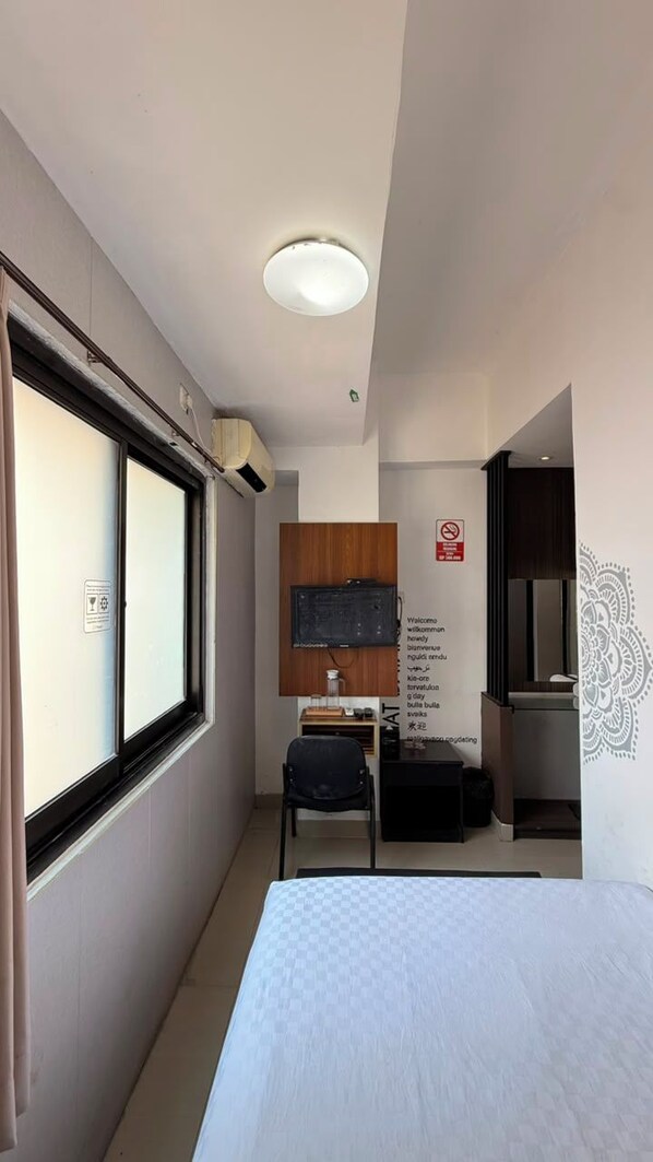 A hotel room with a bed, a desk with a chair, a television, and a window. - De La Charis Hotel (Kediri)
