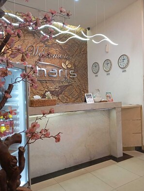 Un mostrador de recepción de hotel con un letrero de bienvenida, relojes que muestran diferentes zonas horarias y un árbol decorativo con flores rosadas.