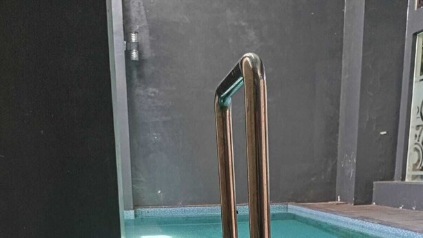 Ein Schwimmbecken mit einer Metallleiter, umgeben von einer dunklen Wand und einer Rasenfläche.