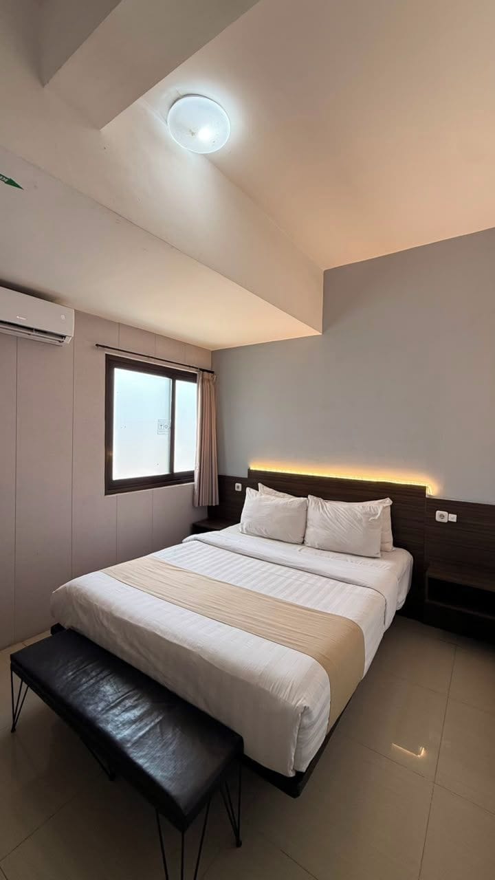 A hotel room with a bed, a bench, a window, and a ceiling light.