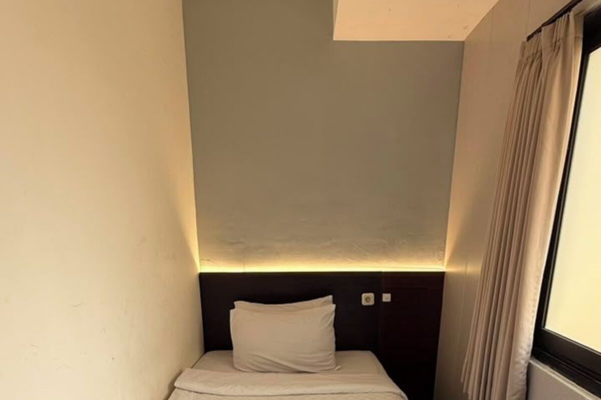 A small, single-bed room with a headboard, a bedside table, and a window with curtains.
