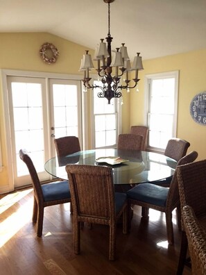 Uma sala de jantar com mesa de vidro, rodeada por cadeiras de vime, um lustre e um relógio na parede.