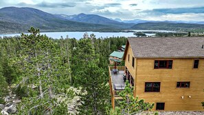A two-story wooden cabin with a deck, surrounded by dense forest and overlooking a lake with mountains in the background. - Spacious Cabin with Foosball Table and Jacuzzi in Grand Lake, Colorado (Grand Lake)