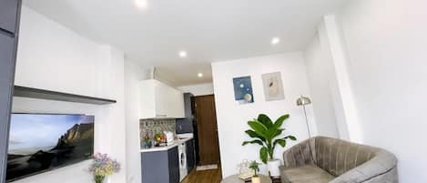 A modern living room with a sofa, a TV, a small table, and a kitchen in the background.