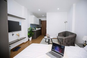 A modern hotel room with a bed, a desk with a laptop, a grey armchair, a small round table with a lamp, a TV mounted on the wall, and a potted plant. - Alaya Lotte Centre Apartment (Ba Dinh)