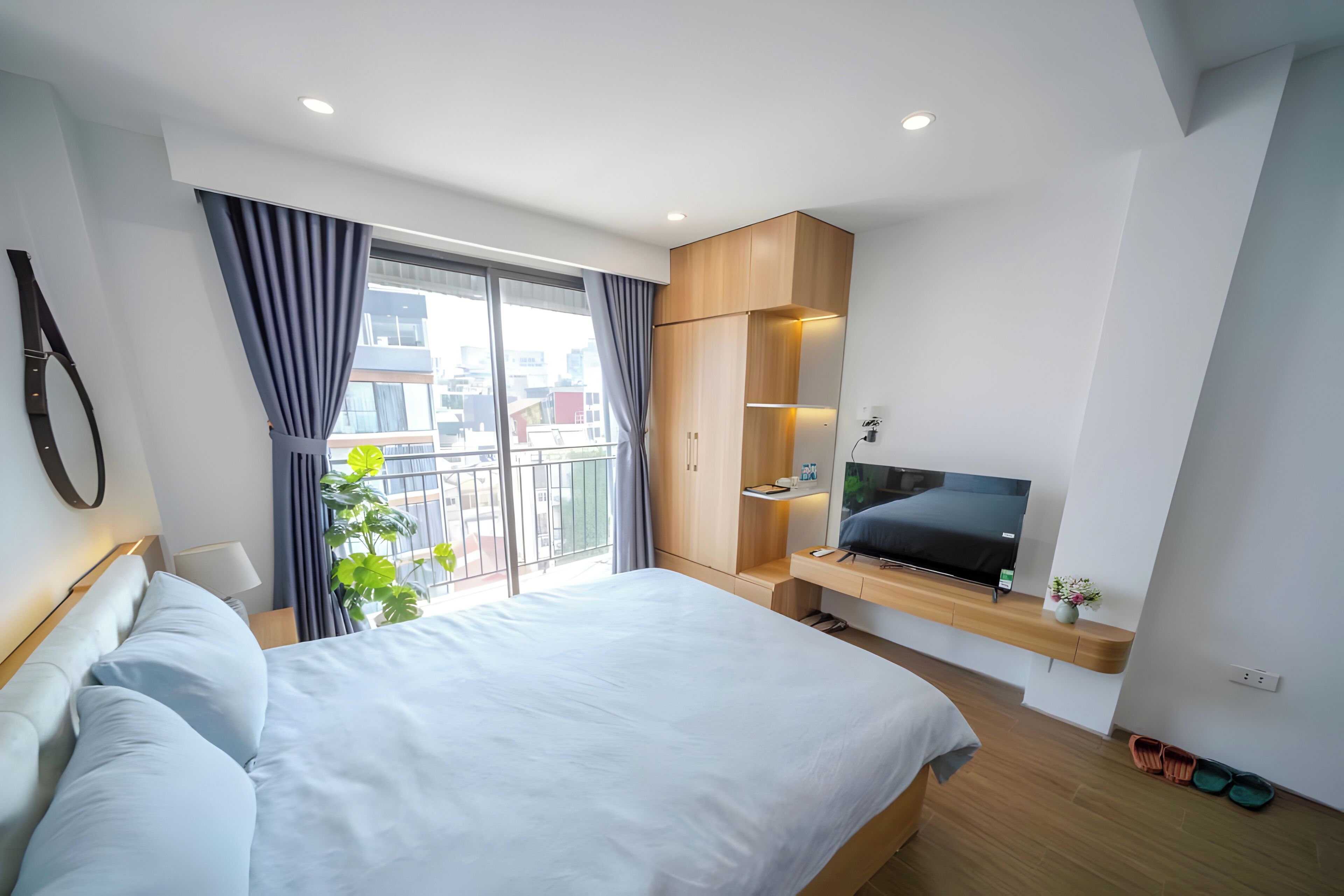 A modern hotel room with a large bed, a TV, a balcony with a plant, and a city view.