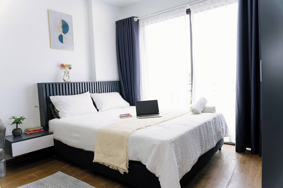Un dormitorio moderno con cama, mesita de noche, una computadora portátil y una ventana con cortinas.
