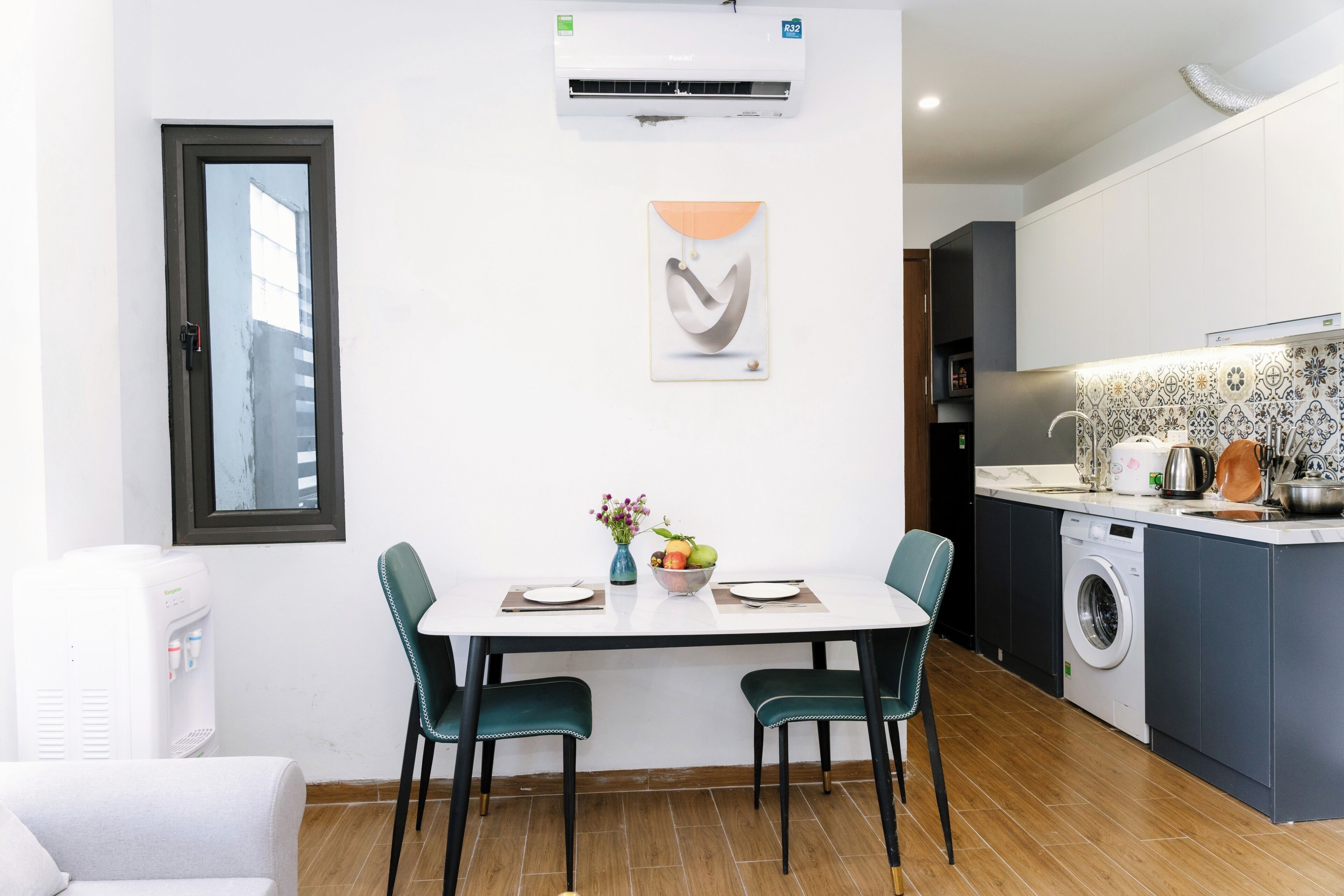 A modern kitchen with a built-in washing machine, a dining area with a white table and teal chairs, and a water dispenser.