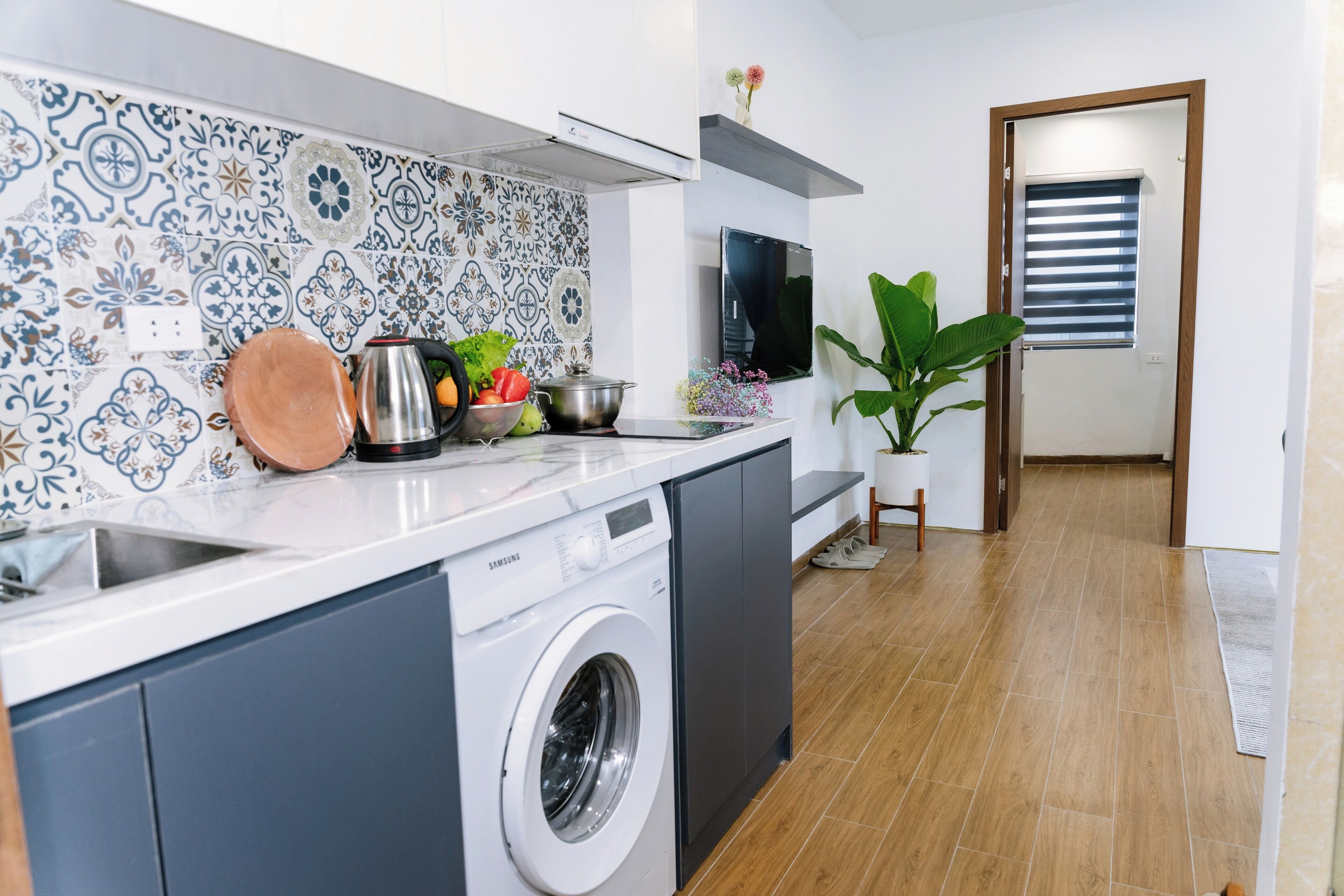 Een moderne keuken met een wasmachine, een spoelbak, een aanrecht met fruit en groenten, een televisie en een grote plant.