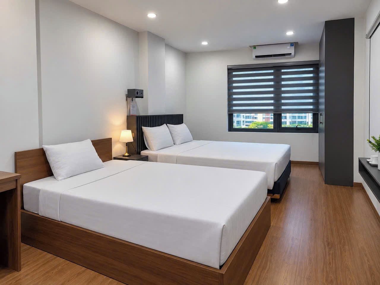 Een hotelkamer met twee bedden, een nachtkastje, een lamp, een raam met zonwering en een airconditioning.