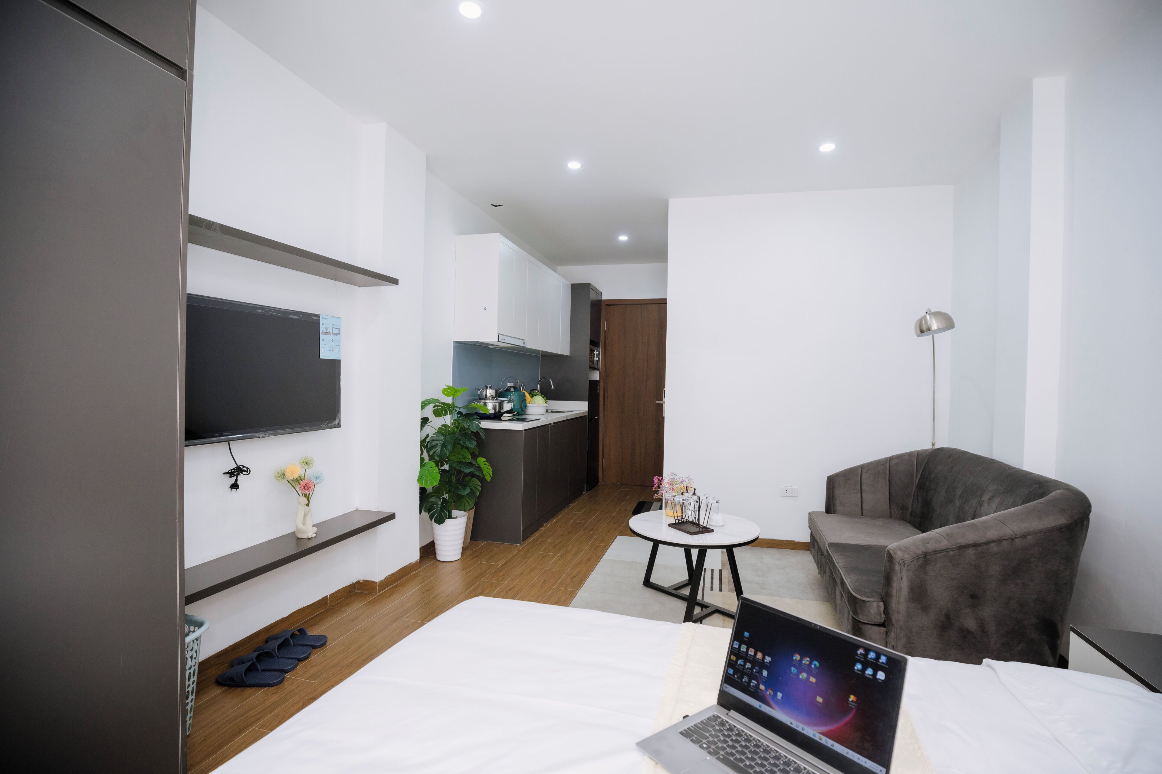 Une chambre d’hôtel moderne avec un lit, un bureau avec un ordinateur portable, un fauteuil gris, une petite table ronde, une plante en pot et, au fond, une kitchenette.
