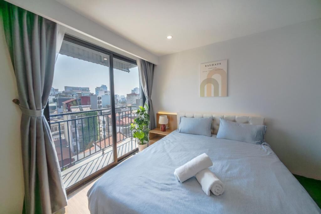 A hotel room with a large bed, a nightstand, a lamp, a view of the city through a balcony, and a framed picture on the wall.