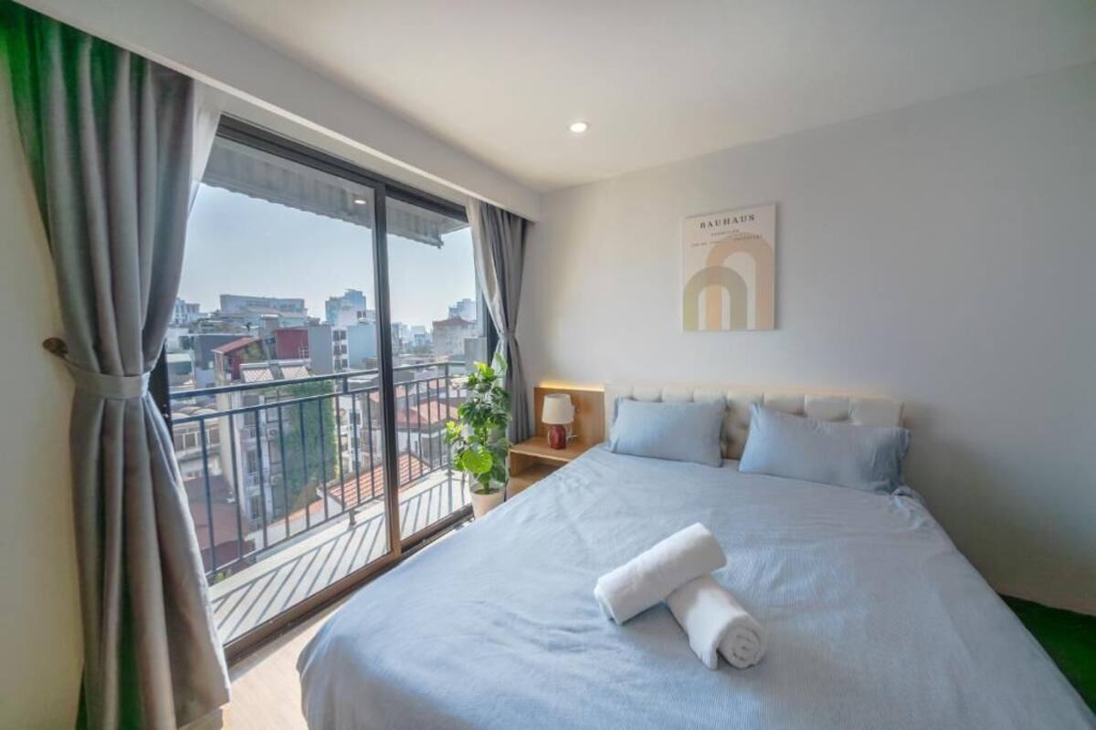 Habitación de hotel con una cama grande, una mesita de noche, una lámpara, vista de la ciudad desde un balcón y un cuadro en la pared.