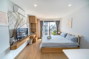 A modern hotel room with a large bed, a desk, a chair, a small table, a plant, and a window with a city view. - Alaya Lotte Centre Apartment (Hanoi)