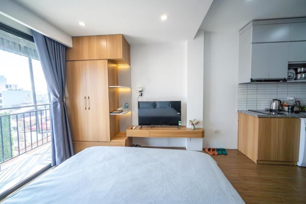 Une chambre d’hôtel moderne équipée d’un lit, d’une télévision, d’une armoire en bois, d’une kitchenette et d’un balcon avec des rideaux.