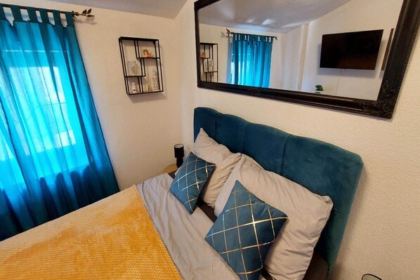 A bedroom with a bed, blue curtains, a bedside table, a mirror, and a television on the wall. - Rooms Argo- Turquoise Double Room (Cres)
