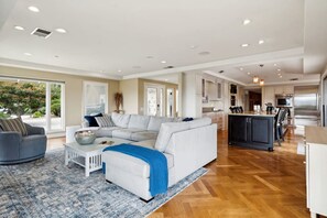 A spacious living room with a large gray sectional sofa, a white coffee table, and a blue throw blanket. The room has a wooden floor and large windows offering a view of the outdoors.
