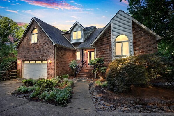A two-story house with a brick exterior, a front garage, and a landscaped garden with various plants and a stone pathway.