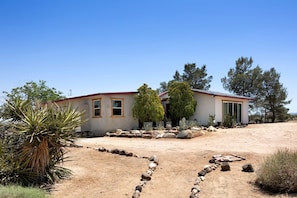 Ein eingeschossiges Haus mit rotem Dach, umgeben von einer trockenen Landschaft mit spärlicher Vegetation und blauem Himmel.