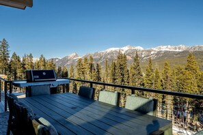 Une terrasse en bois avec une table et des chaises, offrant une vue sur une chaîne de montagnes aux sommets enneigés et une forêt dense.