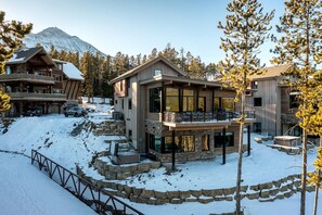 Un chalet moderne à plusieurs étages, doté de fondations en pierre, de grandes fenêtres et d’un balcon offrant une vue sur un paysage enneigé, avec des arbres et une montagne en arrière-plan.