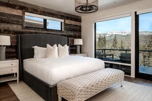 Une chambre à coucher avec un grand lit, un banc, une table de chevet, une lampe et une vue sur les montagnes par une grande fenêtre.