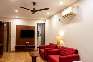 A living room with a ceiling fan, a flat-screen TV mounted on a wooden panel, red upholstered sofas, a small wooden table, and an air conditioning unit on the wall.