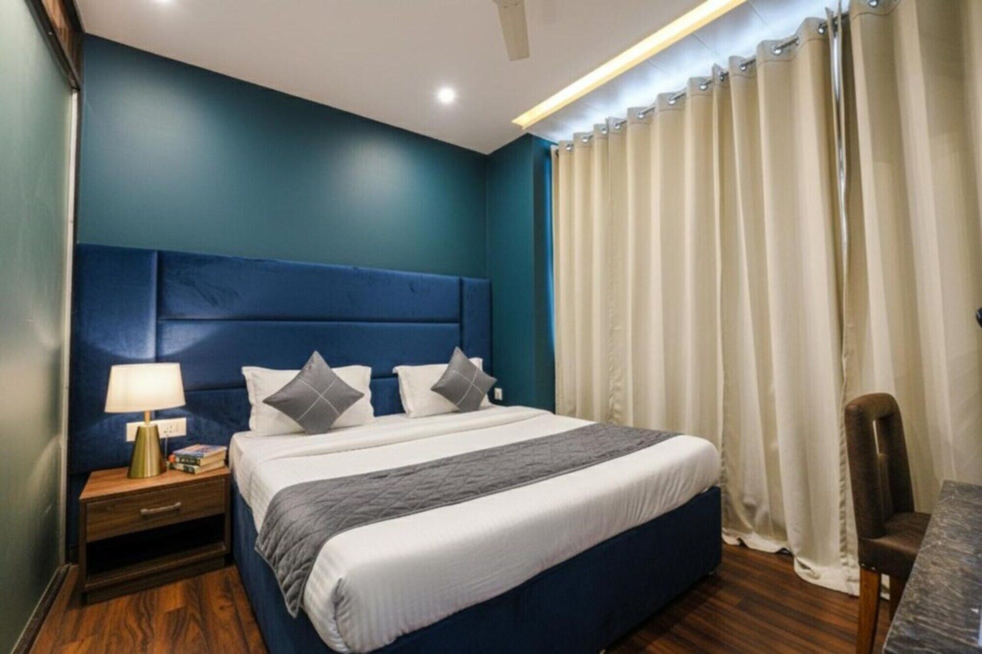 Un dormitorio con cabecera azul, cama con ropa de cama blanca y almohadas grises, mesita de noche con lámpara y una silla.