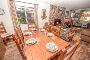 Un coin repas avec une table en bois dressée pour six personnes, entourée de chaises, d’une cheminée en pierre et d’un téléviseur en arrière-plan.