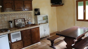 A kitchen with wooden cabinets, a tiled backsplash, a countertop with a coffee maker, a television above the counter, and a window with blinds. - Gîte des Chênes, located in the heart of Béarn. (Arthez-de-Béarn)