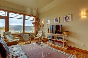 Ein Wohnzimmer mit einer Couch, zwei Sesseln, einem Fernseher, einem Bücherregal und einem großen Fenster mit Blick auf Hügel.