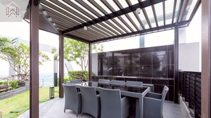 An outdoor patio with a glass-top table and wicker chairs, a pergola overhead, and a view of the sea.