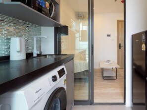 A compact kitchen with a washing machine, a coffee machine, and a small dining area with a table and bench. - Bilik Selesa Tingkat Tinggi dengan Pemandangan Sungai Chao Phraya Mempesonakan (Chang Wat Nonthaburi)