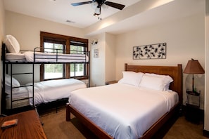 Une chambre avec des lits superposés, un lit simple, un ventilateur de plafond, une fenêtre avec des stores, une lampe fixée au mur et un tableau encadré.