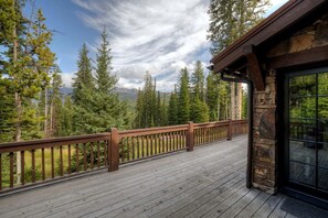 Una terraza de madera con un muro de piedra y puerta de cristal, con vistas a un bosque y montañas.