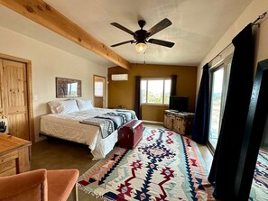 A bedroom with a bed, a desk, a chair, a ceiling fan, and a window with curtains. - Remote Cabin with Hot-Tub and Desert Views in California (Landers)
