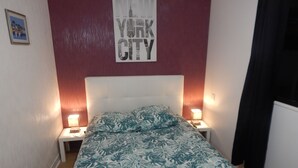 A bedroom with a white headboard, a bed with a tropical leaf pattern bedspread, two bedside tables with lamps, and a wall with a 'NEW YORK CITY' sign. - Beautiful 300 m apartment near the train station and Sillon Beach, within the city walls (Saint-Malo)