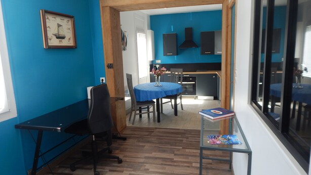 A room with a blue wall, a wooden-framed door, a dining table with chairs, a small table with a book, and a window with blinds. - Beautiful 300 m apartment near the train station and Sillon Beach, within the city walls (Saint-Malo)