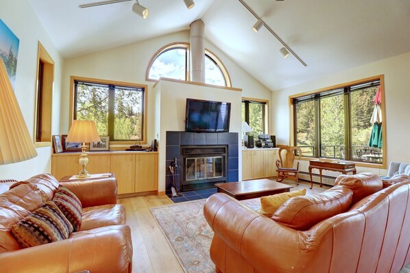 A spacious living room with leather sofas, a fireplace, a television, and large windows offering a view of trees.