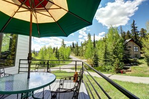 A balcony with a glass table and chairs, an umbrella, and a view of a forested area with a house.