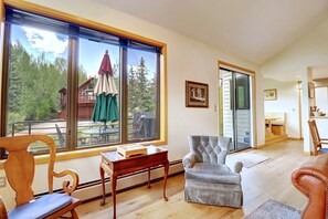 A spacious living room with a large window, a wooden chair, a small table, a patterned armchair, and a view of a cabin and trees.