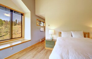 A bedroom with a large window, a bedside table with a lamp, and a bed with white bedding.