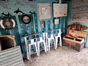 A room with a rustic theme, featuring a bar area with white stools, a vintage trunk, and various wall decorations including a nautical wheel and framed artwork.