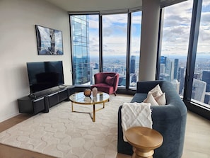 Ein modernes Wohnzimmer mit großem Fernseher, bequemen Sitzmöglichkeiten, einem Glastisch und Blick auf die Stadt durch große Fenster.