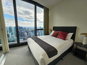 Ein Hotelzimmer mit einem großen Bett, einem Nachttisch, einer Lampe und Blick auf eine Stadtlandschaft durch große Fenster.
