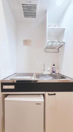 Una kitchenette compacta con un lavamanos, un refrigerador pequeño y una repisa metálica sobre el lavamanos.