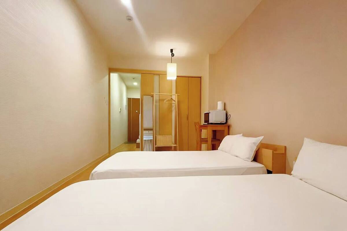 A hotel room with two beds, a wooden wardrobe, a bedside table with a lamp, and a mirror.