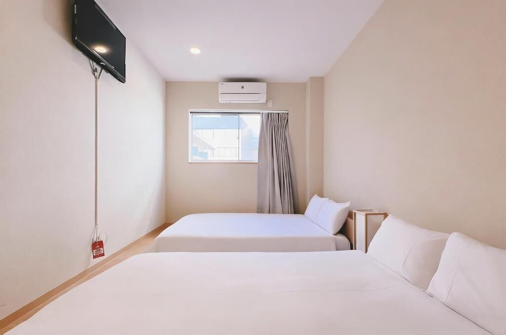 A hotel room with two beds, a TV, an air conditioning unit, and a window with curtains.