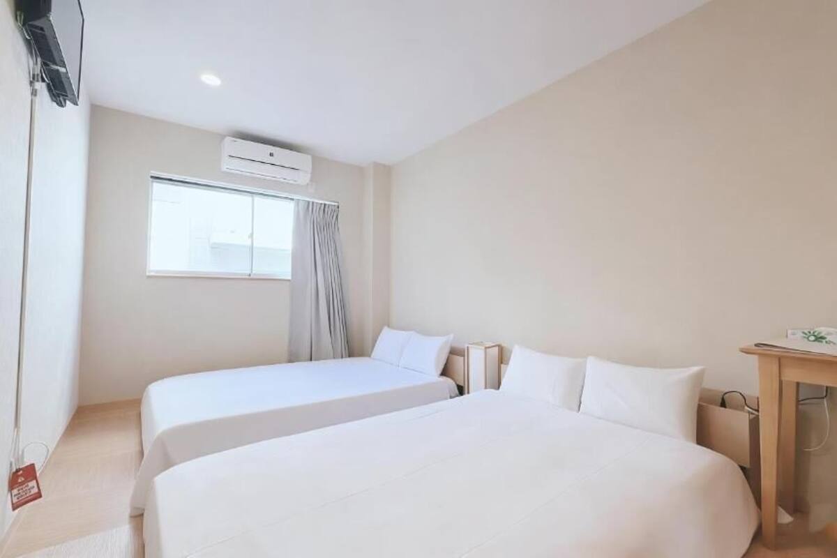 A hotel room with two beds, a desk, a chair, an air conditioning unit, and a window with curtains.