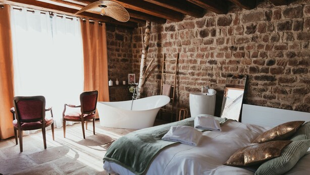 Un dormitorio con una cama grande, una zona de estar con sillones, una bañera y una pared de piedra.